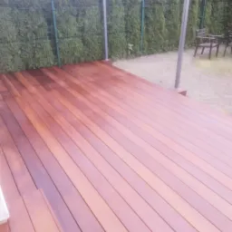 Nowy taras z drewna egzotycznego, deski ułożone równolegle, w tle zielony żywopłot i meble ogrodowe.