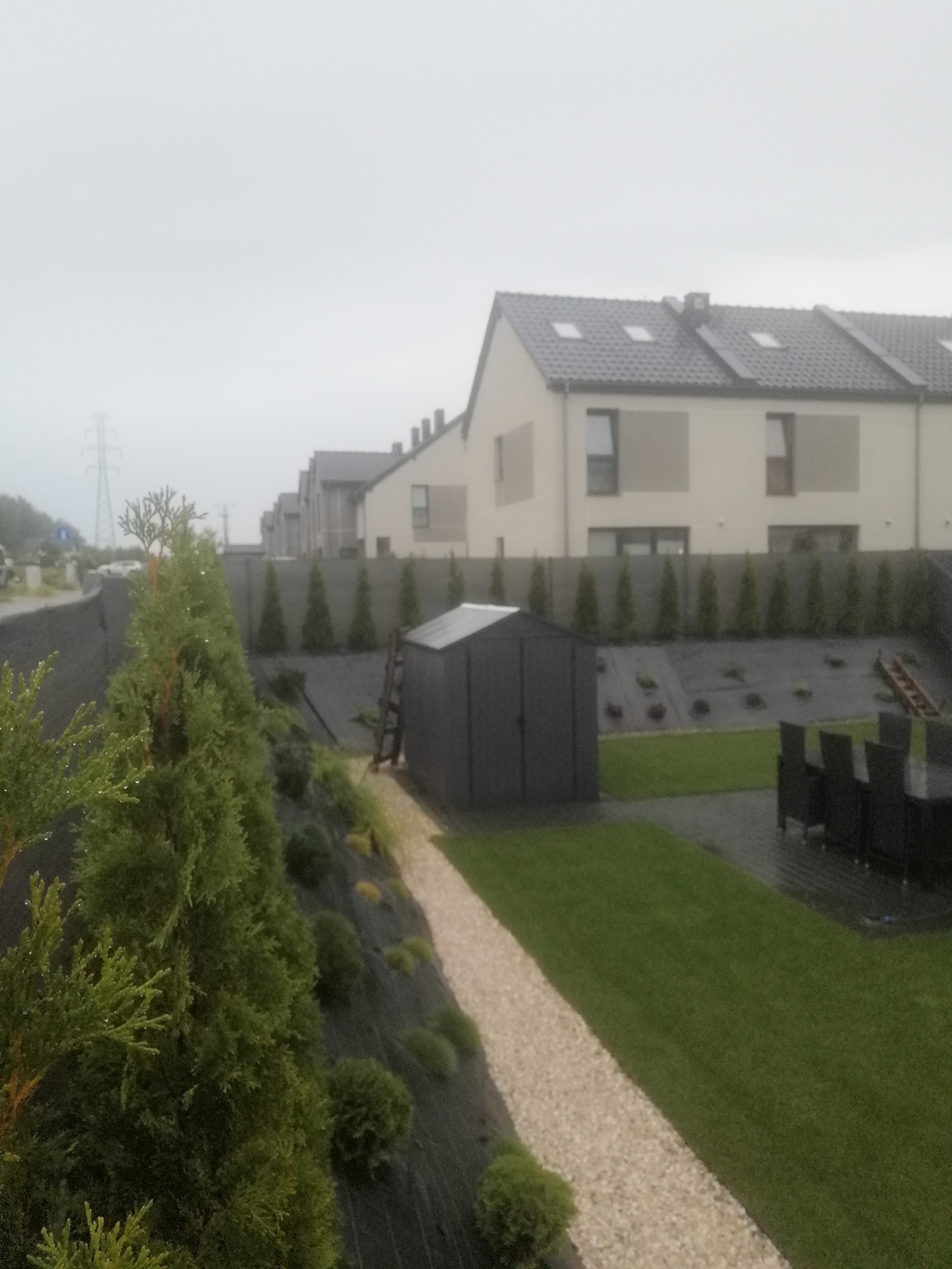 Nowo założony ogród z trawnikiem, ścieżką z jasnych kamieni i podwyższoną rabatą obsadzoną tujami wzdłuż ogrodzenia.