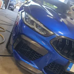 Niebieski sportowy samochód, widok z przodu, z elementami karbonowymi na zderzaku, zaparkowany w garażu.