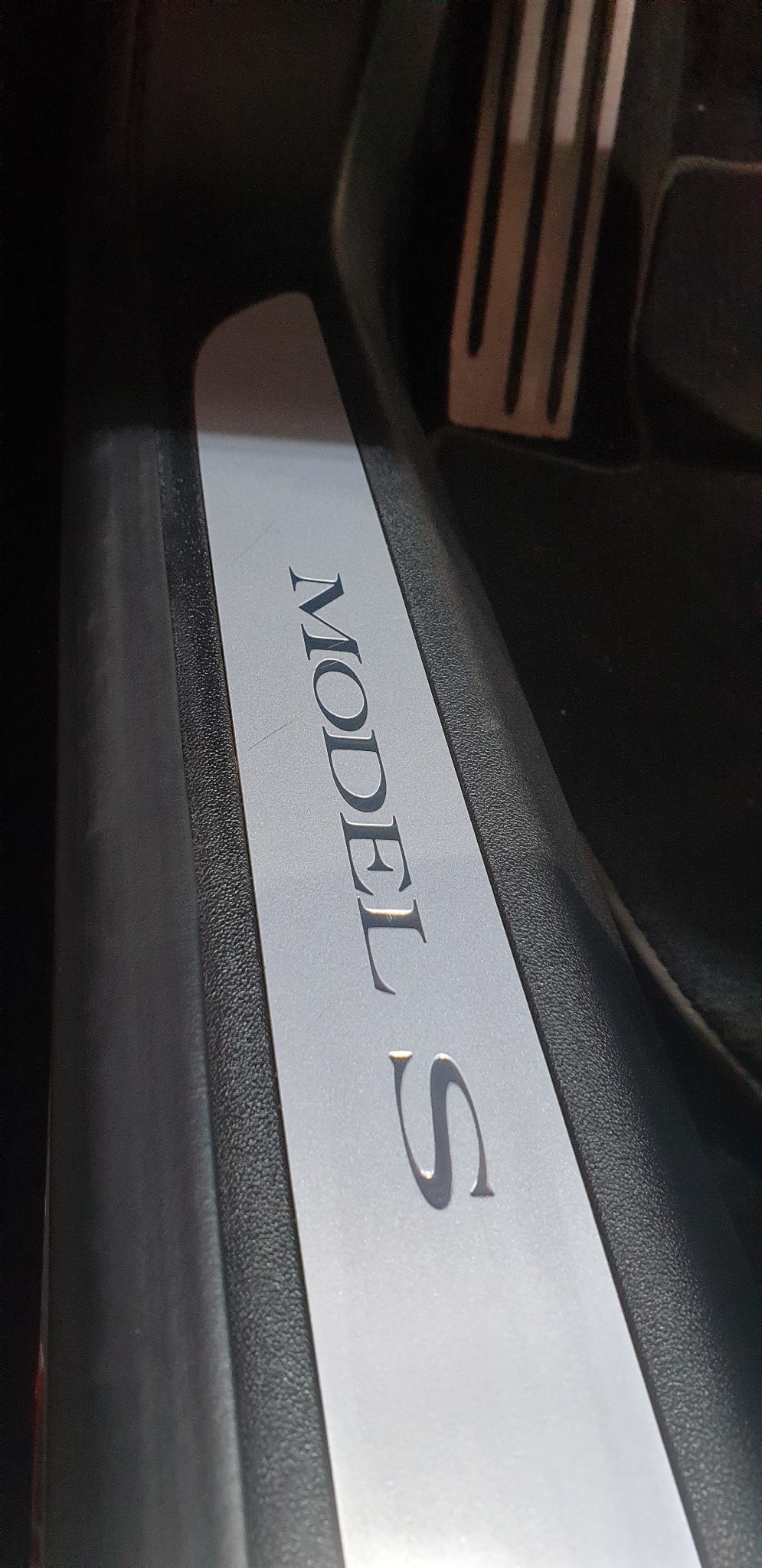 Wnętrze samochodu Tesla Model S, widoczna listwa progowa z napisem 'MODEL S' w srebrnym kolorze, umieszczona na czarnym tle.
