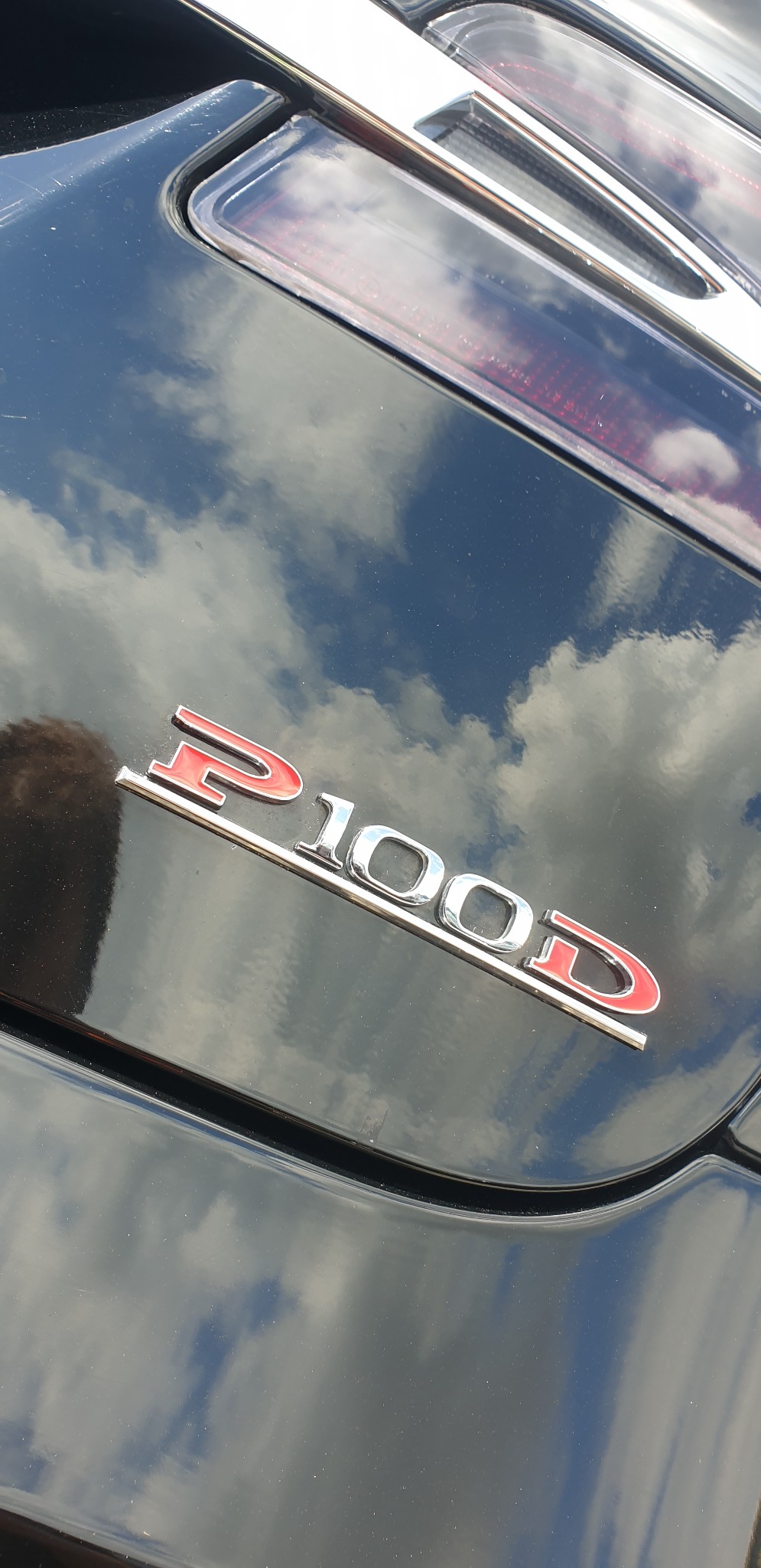 Błyszcząca, czarna karoseria samochodu z chromowanym emblematem P100D odbijającym pochmurne niebo.