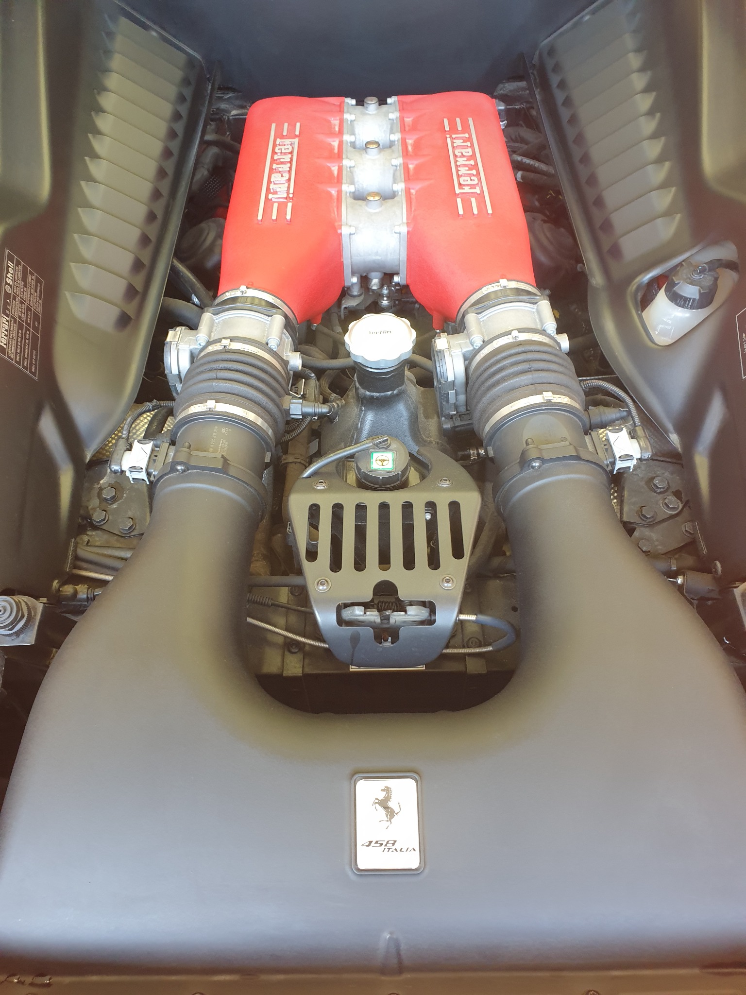 Widok z góry na silnik Ferrari 458 Italia, z czerwonymi pokrywami głowic i czarną obudową filtra powietrza, ukazujący detale konstrukcyjne.