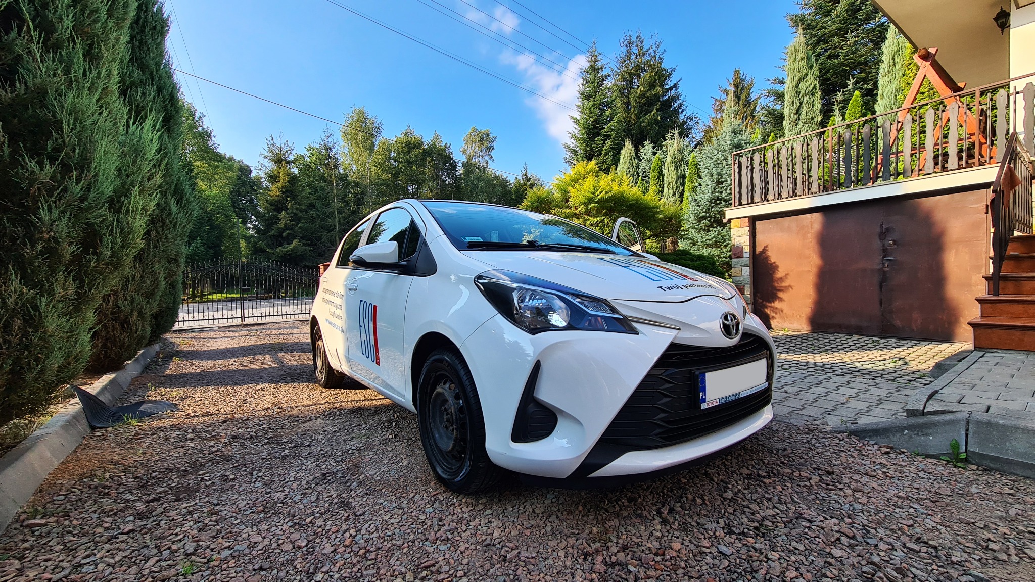 Biała Toyota Yaris z logo firmy na tle zieleni i błękitnego nieba, zaparkowana na żwirowym podjeździe przed domem.