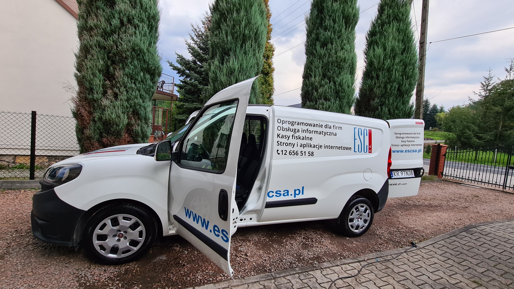 Biały samochód dostawczy z otwartymi drzwiami, oklejony reklamą firmy oferującej oprogramowanie dla firm, obsługę informatyczną, kasy fiskalne oraz strony i aplikacje internetowe, zaparkowany...