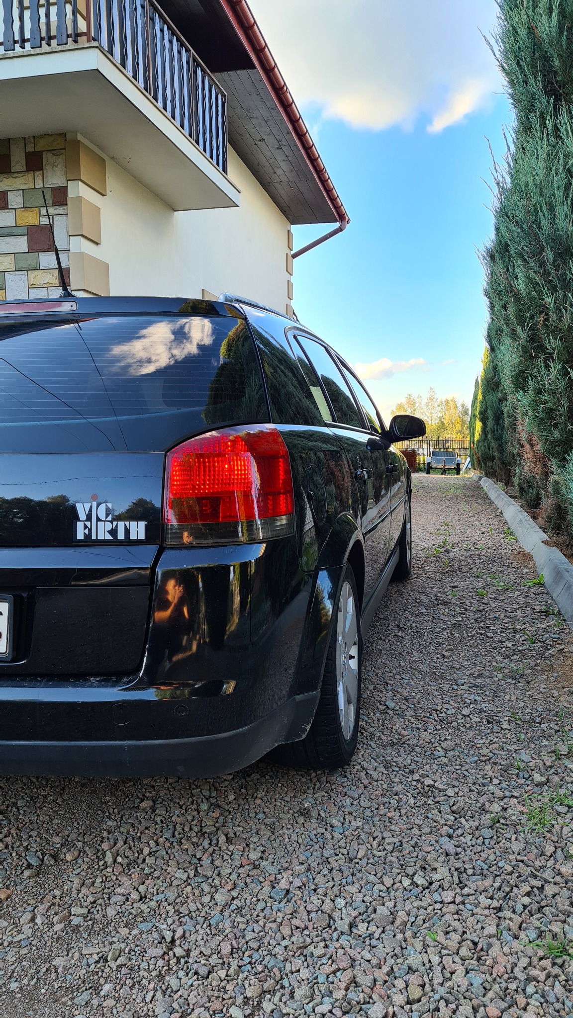Czarne kombi zaparkowane na żwirowym podjeździe obok żywopłotu, z naklejką 'Vic Firth' na tylnej szybie.