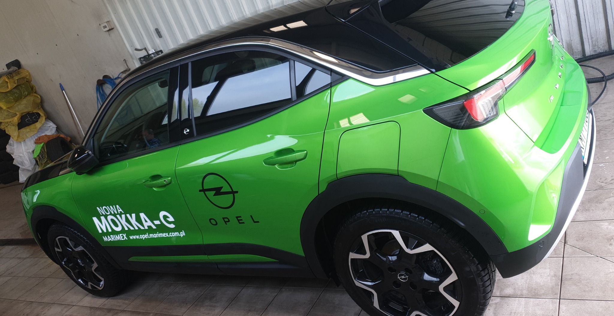 Świeżo umyty, jaskrawozielony Opel Mokka-e z czarnym dachem, zaparkowany w myjni, widok z tyłu i boku, z widocznymi kroplami wody na karoserii.
