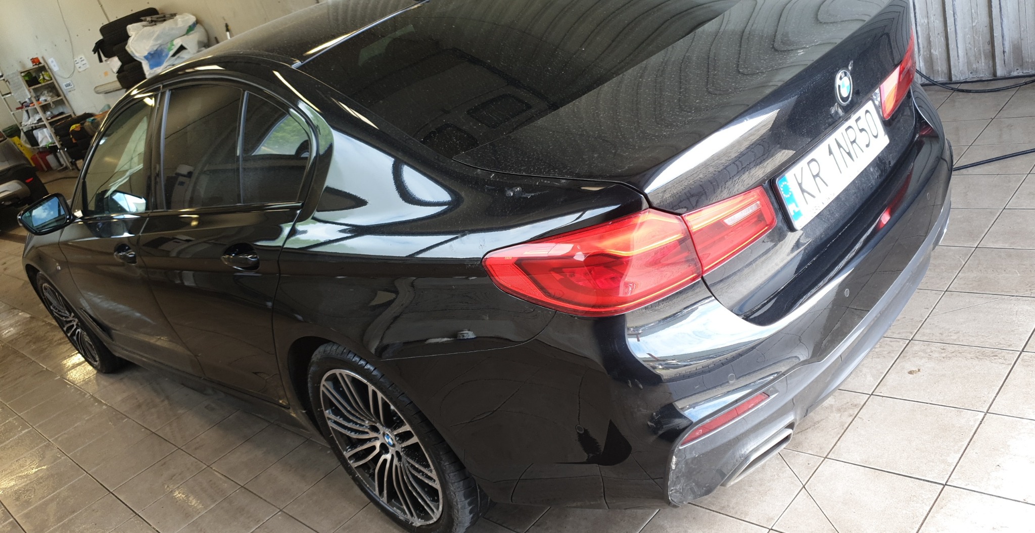 Czarny samochód BMW sedan, widoczny z tyłu pod kątem, z widocznym numerem rejestracyjnym, lekko zakurzony, stojący na kafelkach w pomieszczeniu przypominającym myjnię.