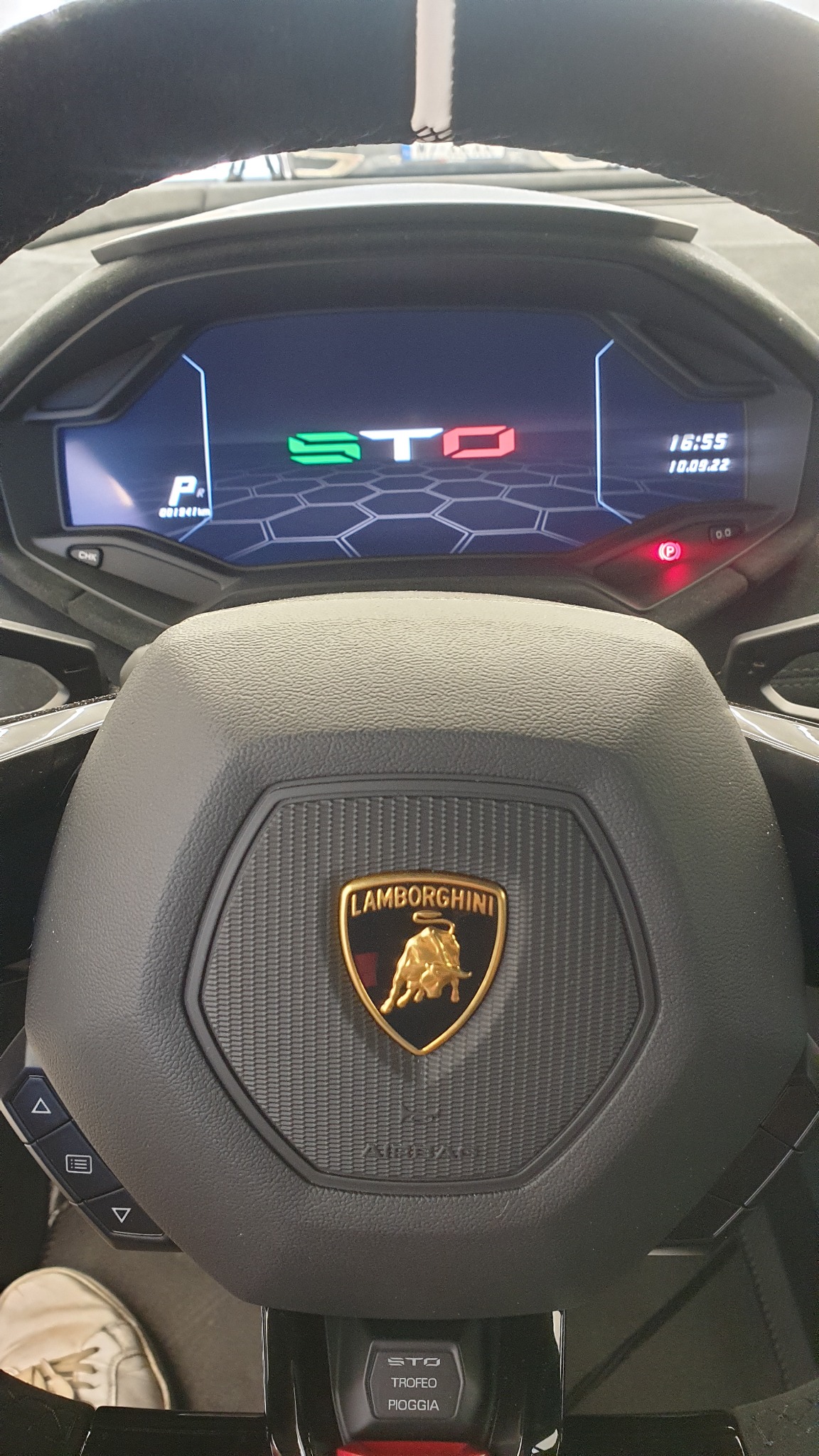 Wnętrze Lamborghini STO: kierownica z logo, cyfrowy wyświetlacz z informacjami o pojeździe, widoczny fragment białego buta kierowcy.