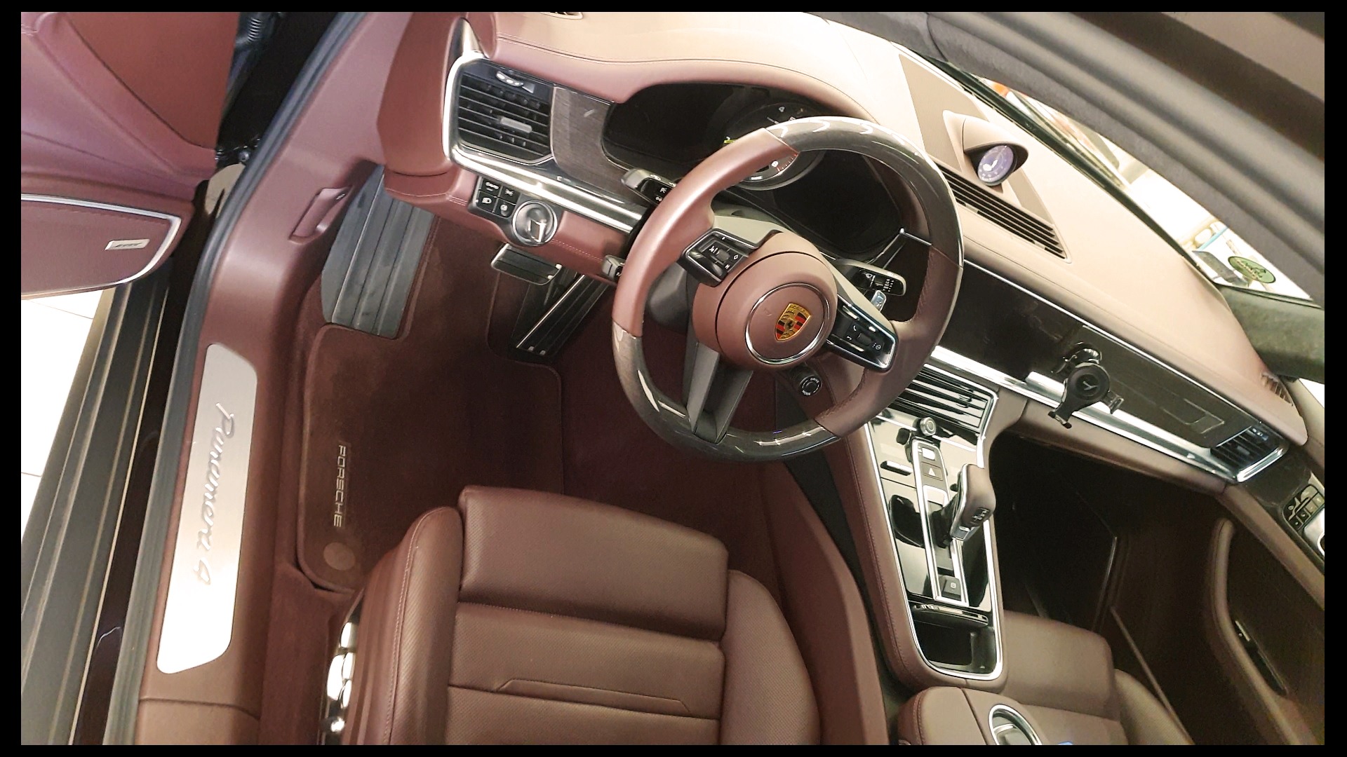 Wnętrze samochodu Porsche Panamera z brązową skórzaną tapicerką, widoczna kierownica z logo Porsche, deska rozdzielcza i fotel kierowcy, z otwartymi drzwiami po lewej stronie.