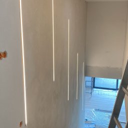 Jar-Mich jarosław Sokołowski Michał Niedźwiecki s.c. - Montaż pionowych, wpuszczanych opraw oświetleniowych LED w surowej ścianie klatki schodowej, widoczna aluminiowa drabina i instalacja elektryczna w puszkach.
