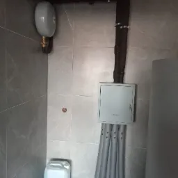 Szare pomieszczenie techniczne z instalacją rur w izolacji, zbiornikiem wyrównawczym, skrzynką elektryczną Prawtech Geothermal i kanistrem z płynem na podłodze.