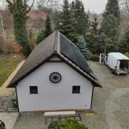 Jar-Mich jarosław Sokołowski Michał Niedźwiecki s.c. - Widok z góry na budynek z zamontowanymi panelami fotowoltaicznymi na dachu, dekoracyjnym kołem na ścianie i zaparkowanym białym vanem z otwartymi drzwiami w tle.