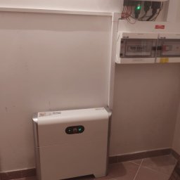 Jar-Mich jarosław Sokołowski Michał Niedźwiecki s.c. - Biały magazyn energii Huawei z podłączonymi przewodami i skrzynką rozdzielczą na ścianie wewnątrz budynku, widoczne płytki podłogowe.