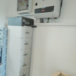 Jar-Mich jarosław Sokołowski Michał Niedźwiecki s.c. - Biały falownik Fronius SYMO i bateria BYD LVL 15.4 w pomieszczeniu, z widoczną skrzynką zabezpieczeń i fragmentem ramy transportowej