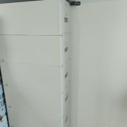 Jar-Mich jarosław Sokołowski Michał Niedźwiecki s.c. - Biały, modułowy magazyn energii ułożony pionowo, zainstalowany w Zgorzelcu przy białej ścianie. Widoczne czarne elementy na górze urządzenia i przewody elektryczne przy suficie.