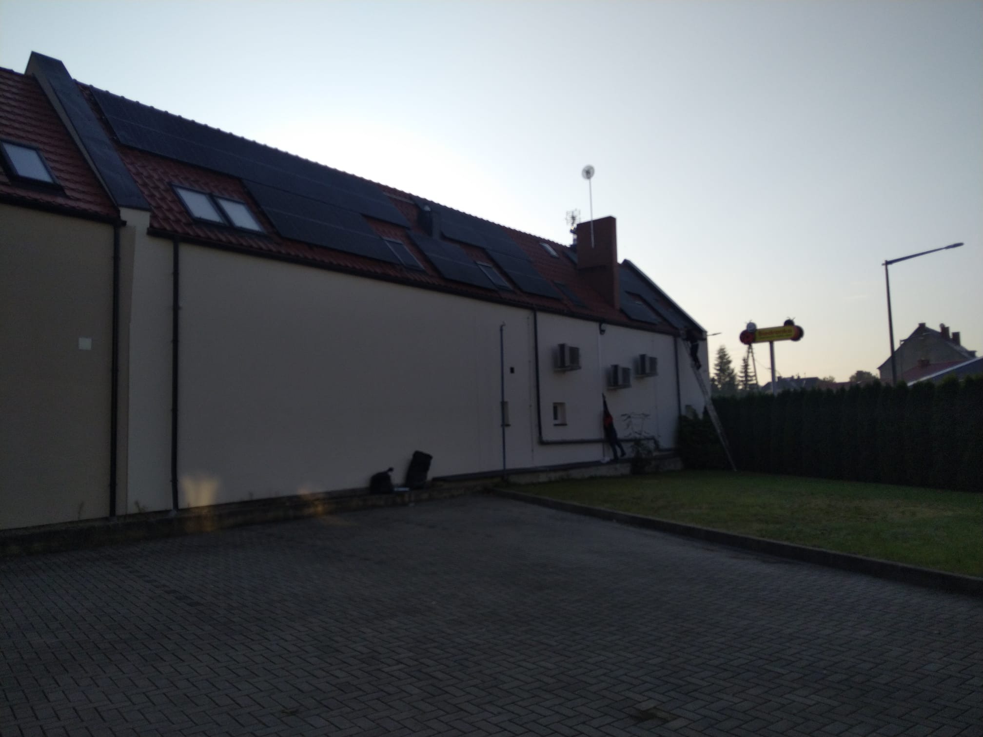 Dwupiętrowy budynek z panelami słonecznymi na dachu i kilkoma jednostkami klimatyzacji na ścianie, widziany z parkingu wyłożonego kostką brukową.