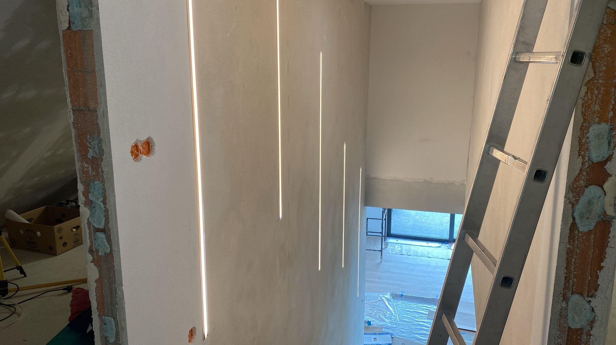 Montaż pionowych, wpuszczanych opraw oświetleniowych LED w surowej ścianie klatki schodowej, widoczna aluminiowa drabina i instalacja elektryczna w puszkach.
