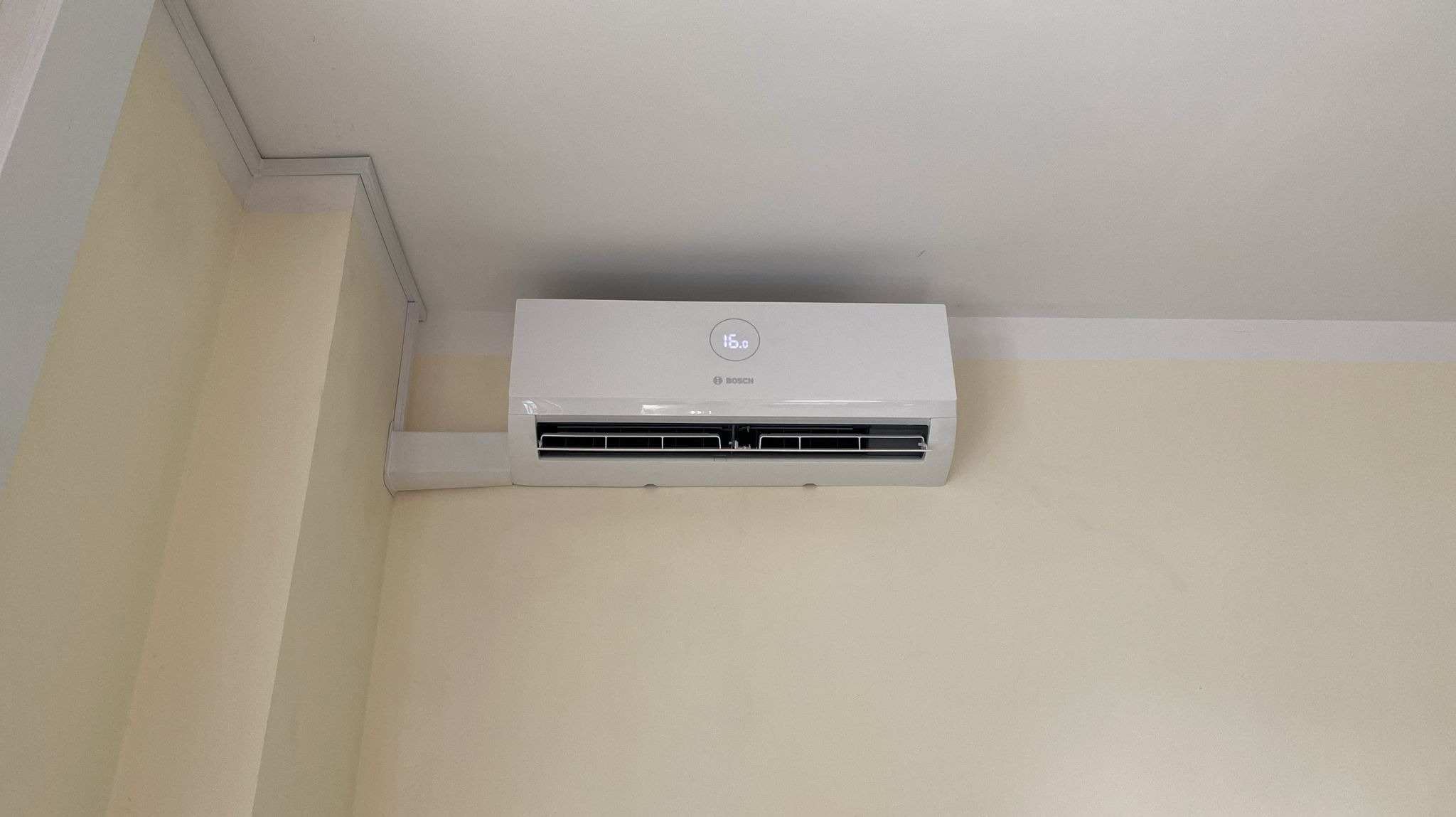 Biała jednostka wewnętrzna klimatyzacji marki Bosch zamontowana na kremowej ścianie, widoczny wyświetlacz temperatury 16 stopni Celsjusza.
