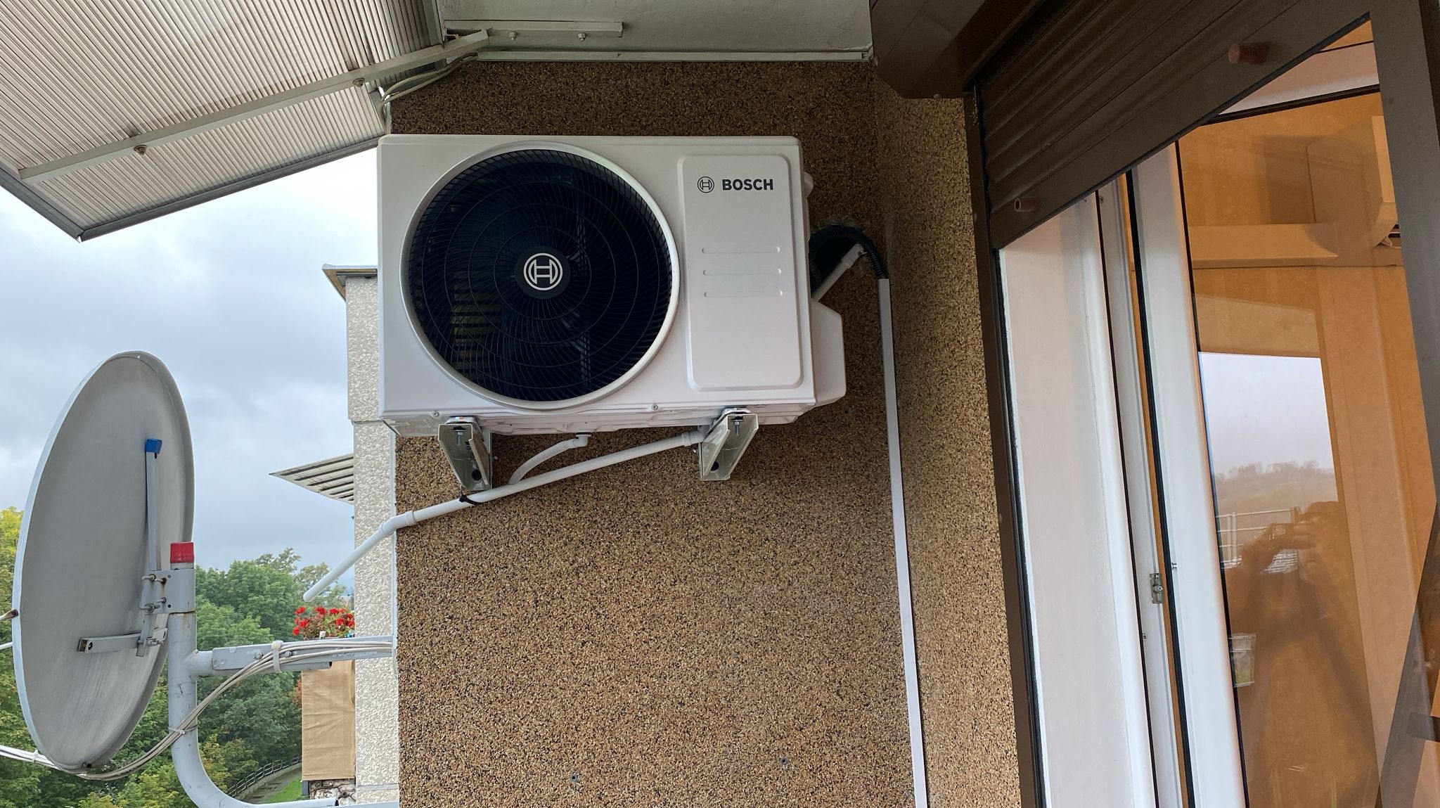 Biała jednostka zewnętrzna z logo Bosch zamontowana na balkonie, widoczna antena satelitarna i częściowo opuszczone rolety okienne.