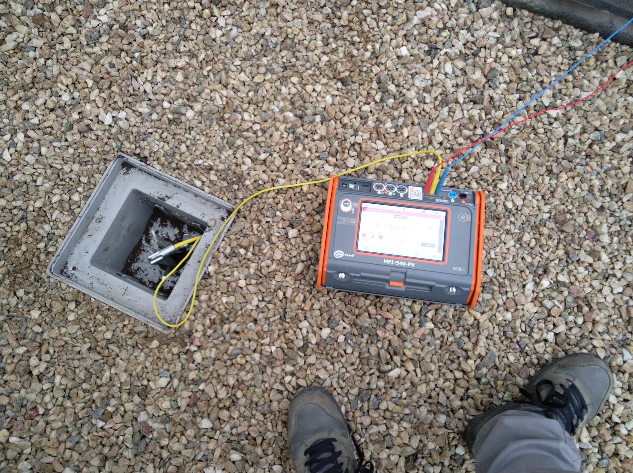 Miernik MPI-540-PV podłączony do instalacji w skrzynce przyłączeniowej na tle żwirowego dachu, obok widoczna stopa w szarym bucie.