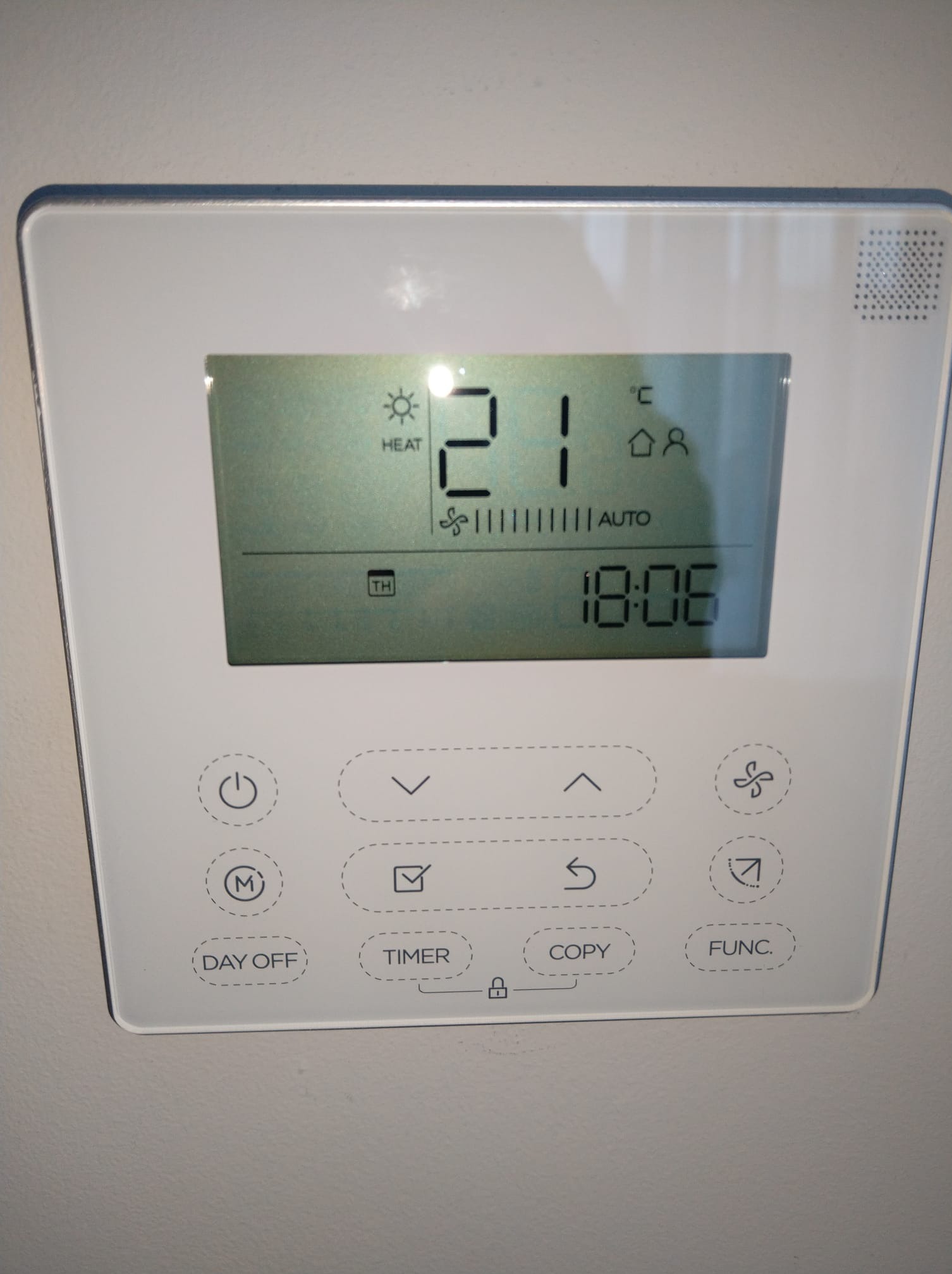 Biały panel sterowania klimatyzacją z wyświetlaczem pokazującym temperaturę 21 stopni Celsjusza, tryb grzania i godzinę 18:06, zamontowany na ścianie.