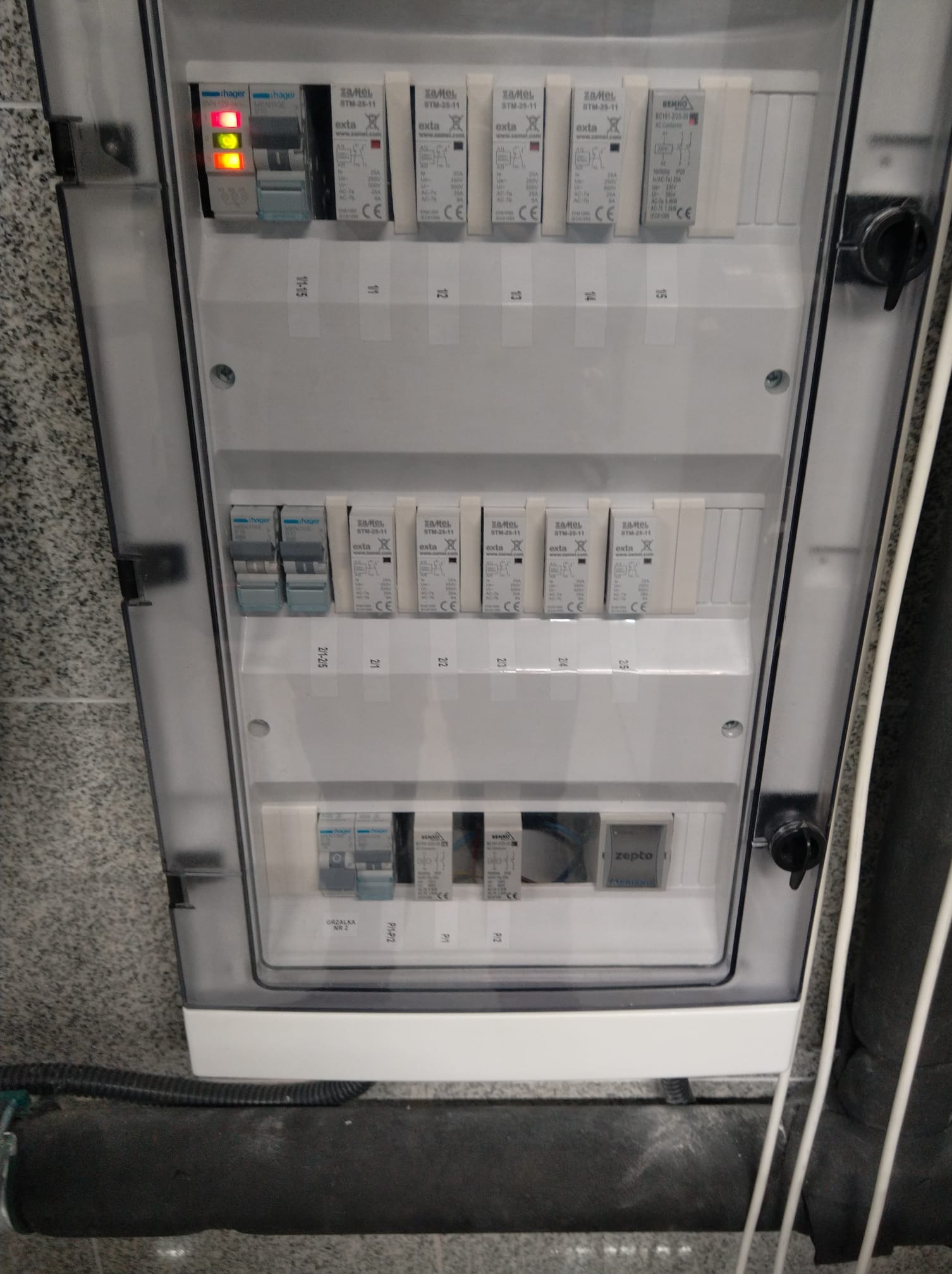 Rozdzielnia elektryczna z bezpiecznikami i sygnalizacją LED, zamontowana na ścianie z szarym tłem, widoczne rury instalacyjne.