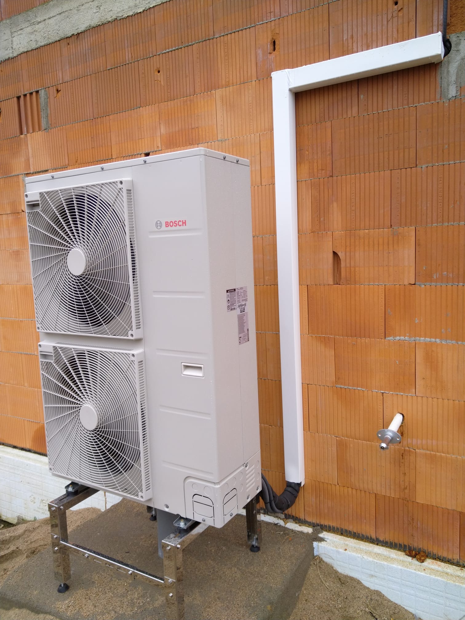 Zewnętrzna jednostka pompy ciepła Bosch zamontowana na metalowym stelażu przy ścianie z cegły ceramicznej z widocznymi rurami odprowadzającymi skropliny.