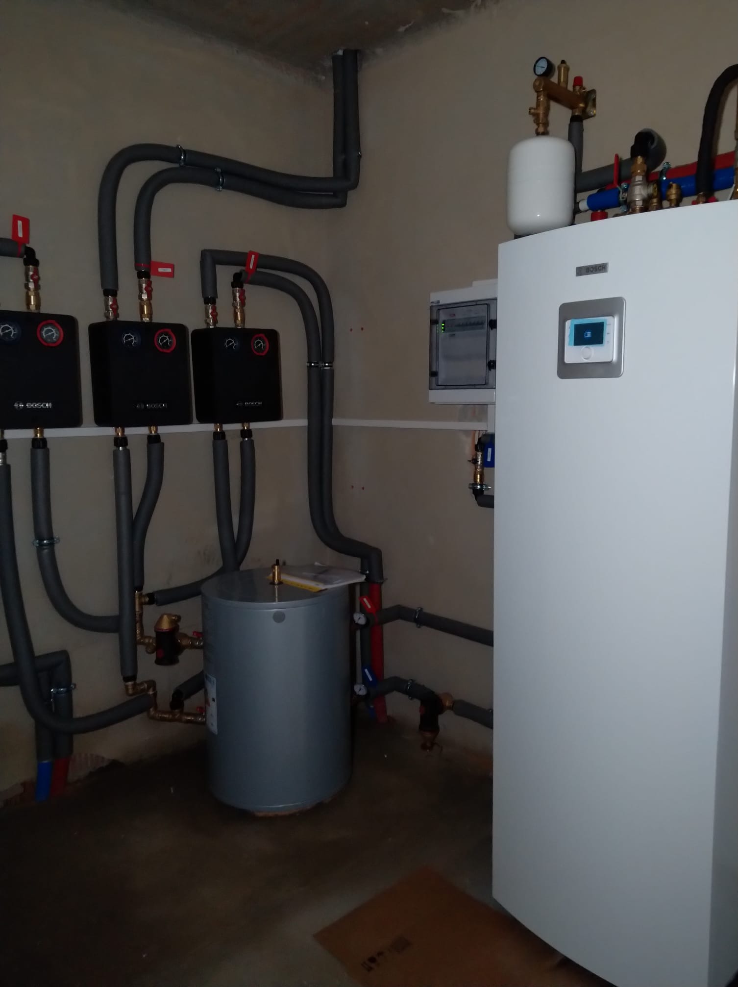 Instalacja z białym zbiornikiem buforowym, trzema czarnymi modułami sterującymi Bosch i szarymi rurami w pomieszczeniu technicznym.