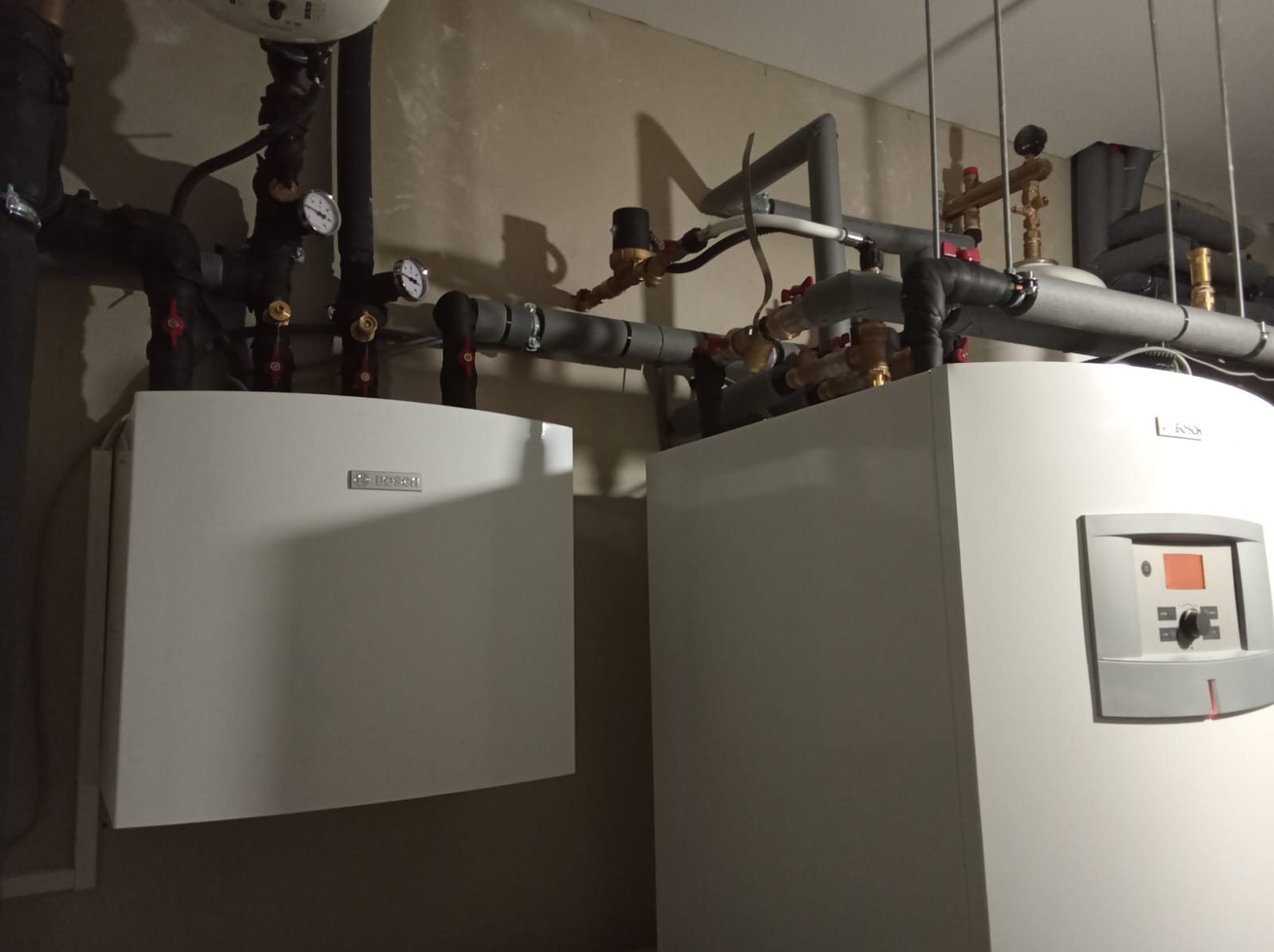 Dwa białe urządzenia grzewcze marki Bosch, z widocznymi rurami, zaworami i manometrami, zamontowane na szarej ścianie.
