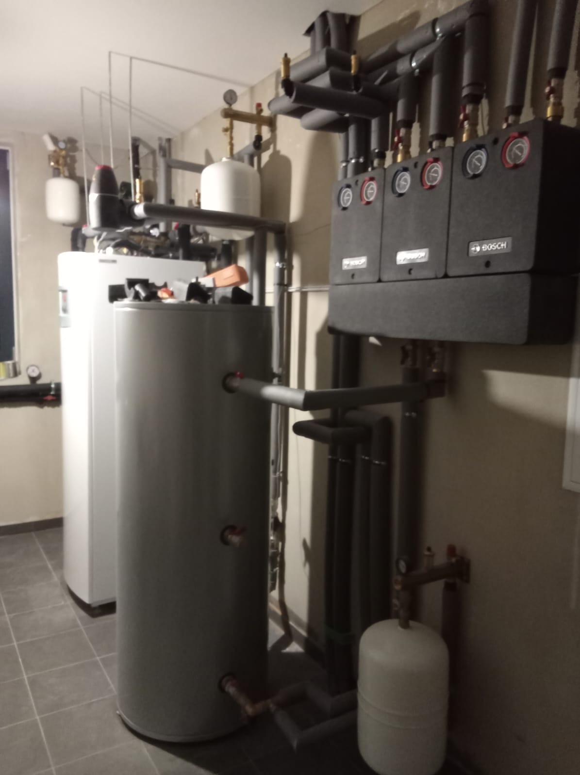 Szara instalacja grzewcza z trzema czarnymi modułami Bosch, rurami w izolacji i zbiornikami w pomieszczeniu technicznym, widoczne manometry i zawory.