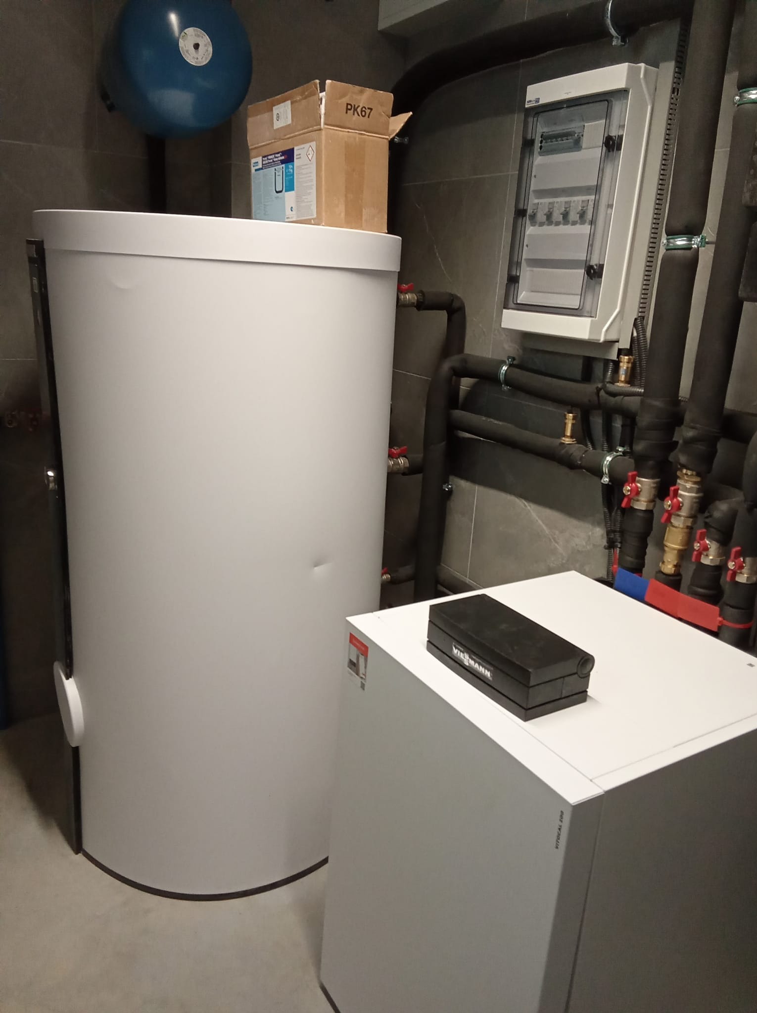Instalacja kotłowni z białym zasobnikiem ciepłej wody i kotłem grzewczym Viessmann Vitocal 200, widoczne rury w izolacji, skrzynka elektryczna oraz niebieski zbiornik wyrównawczy.