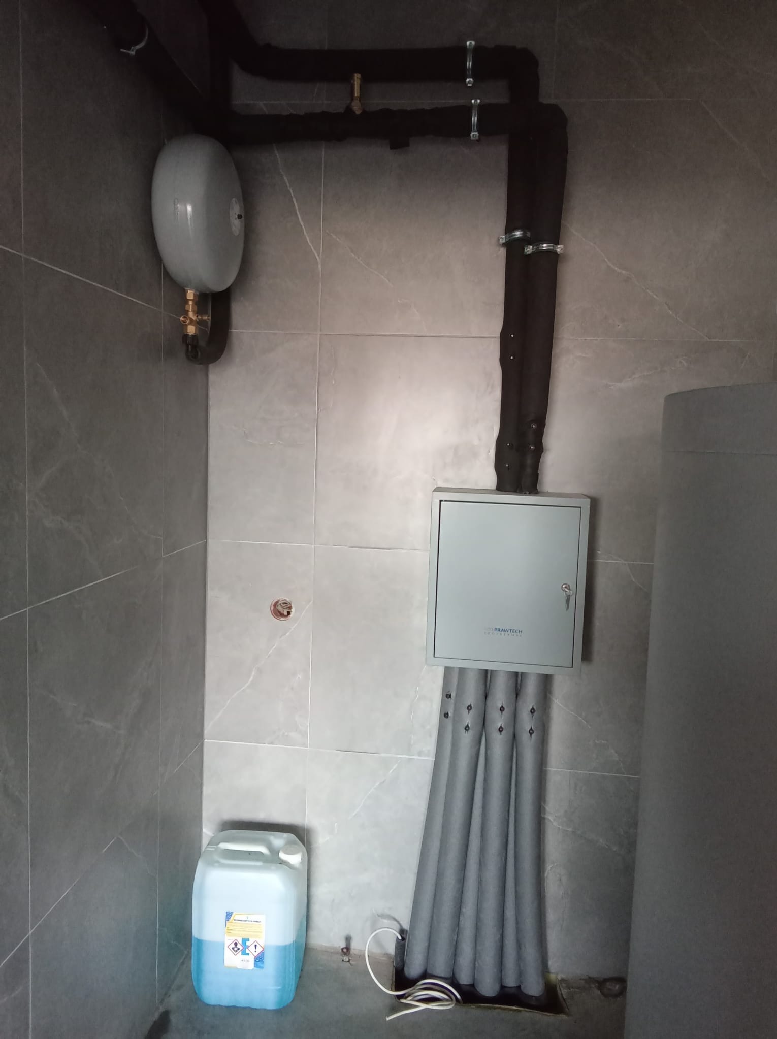 Szare pomieszczenie techniczne z instalacją rur w izolacji, zbiornikiem wyrównawczym, skrzynką elektryczną Prawtech Geothermal i kanistrem z płynem na podłodze.