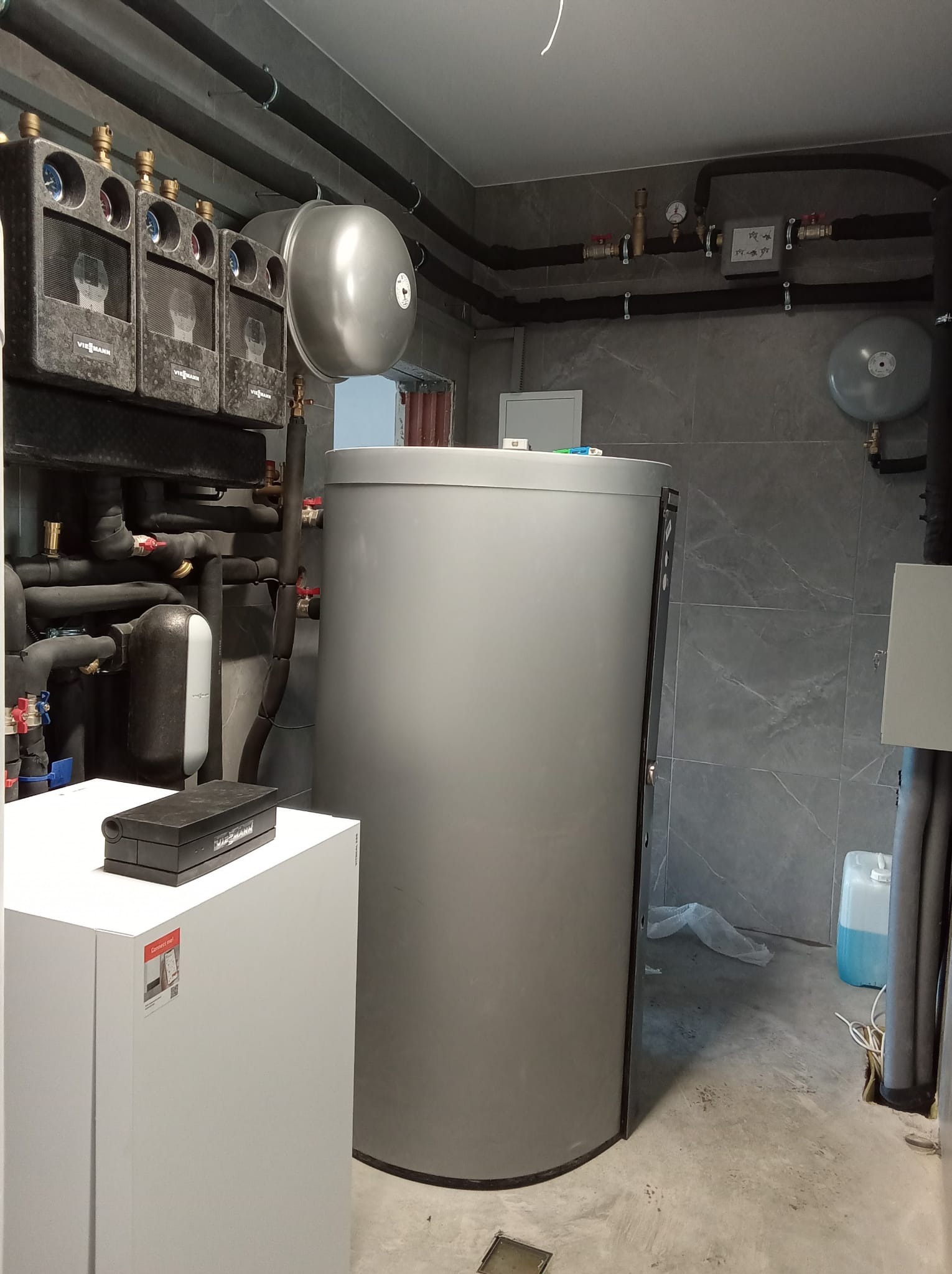 Szara kotłownia z instalacją grzewczą, zbiornikiem buforowym i orurowaniem pod sufitem, widoczne elementy marki Viessmann.