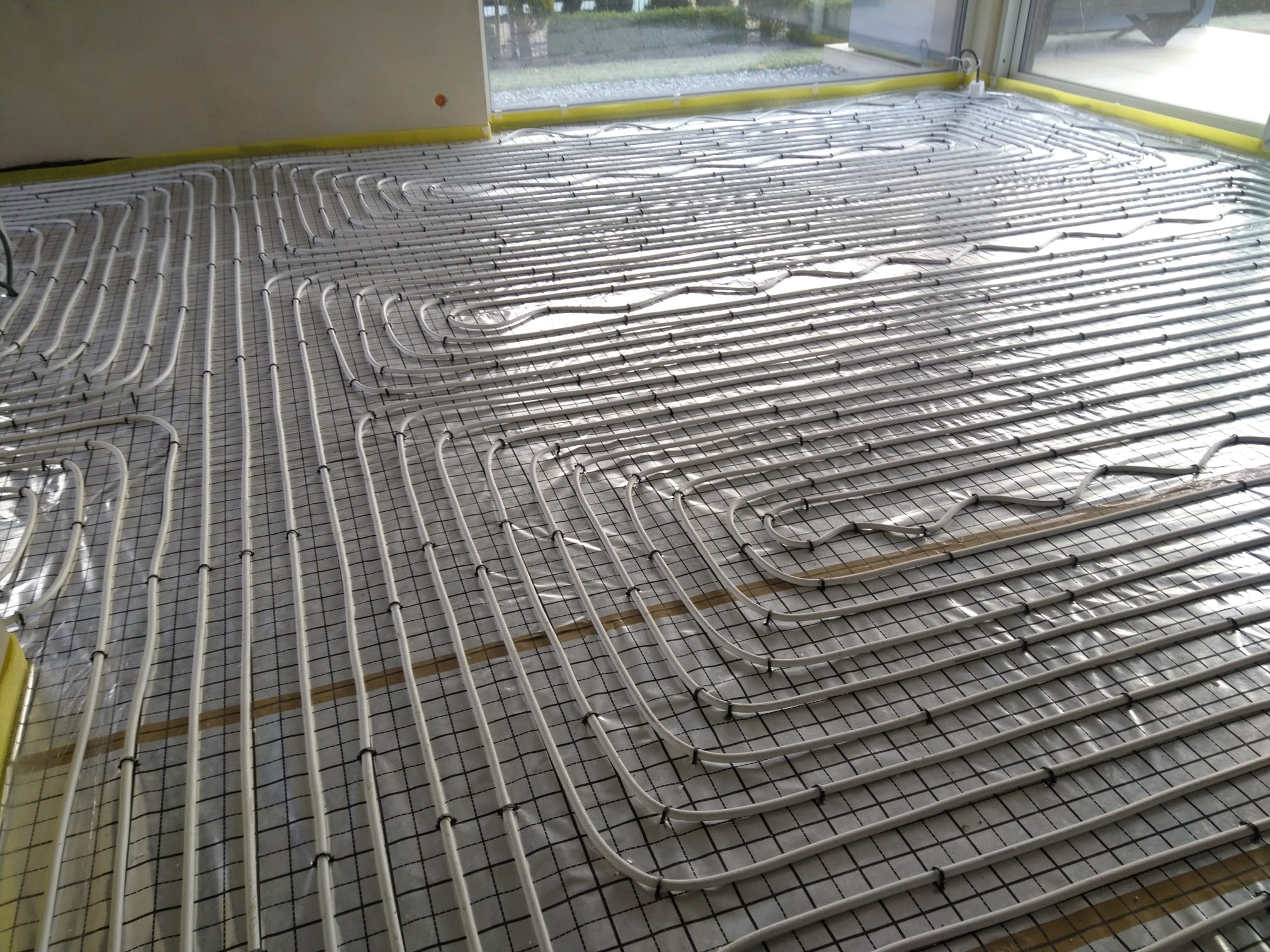 Rozłożona instalacja ogrzewania podłogowego z białych rurek na czarnej siatce, widoczne okna w tle.