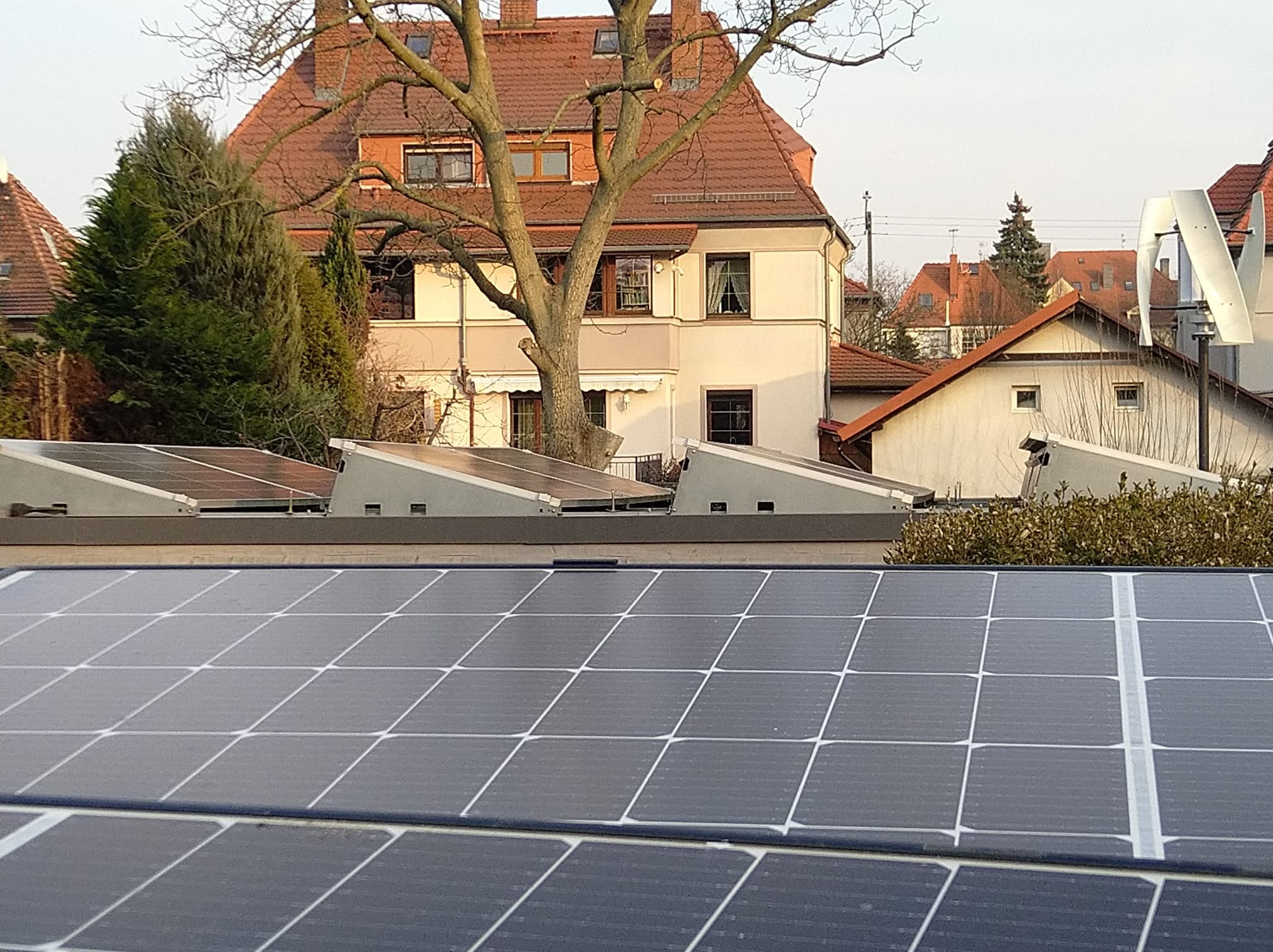 Panel słoneczny z widocznymi liniami podziału ogniw, zamontowany na dachu budynku mieszkalnego w tle zabudowa jednorodzinna oraz turbina wiatrowa.