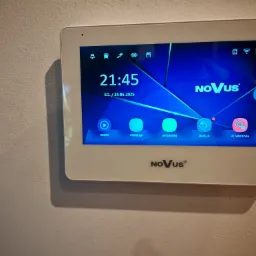 Biały panel dotykowy wideodomofonu Novus zamontowany na ścianie, wyświetlający menu z ikonami i aktualną godziną. Obok widoczny fragment szklanego włącznika.