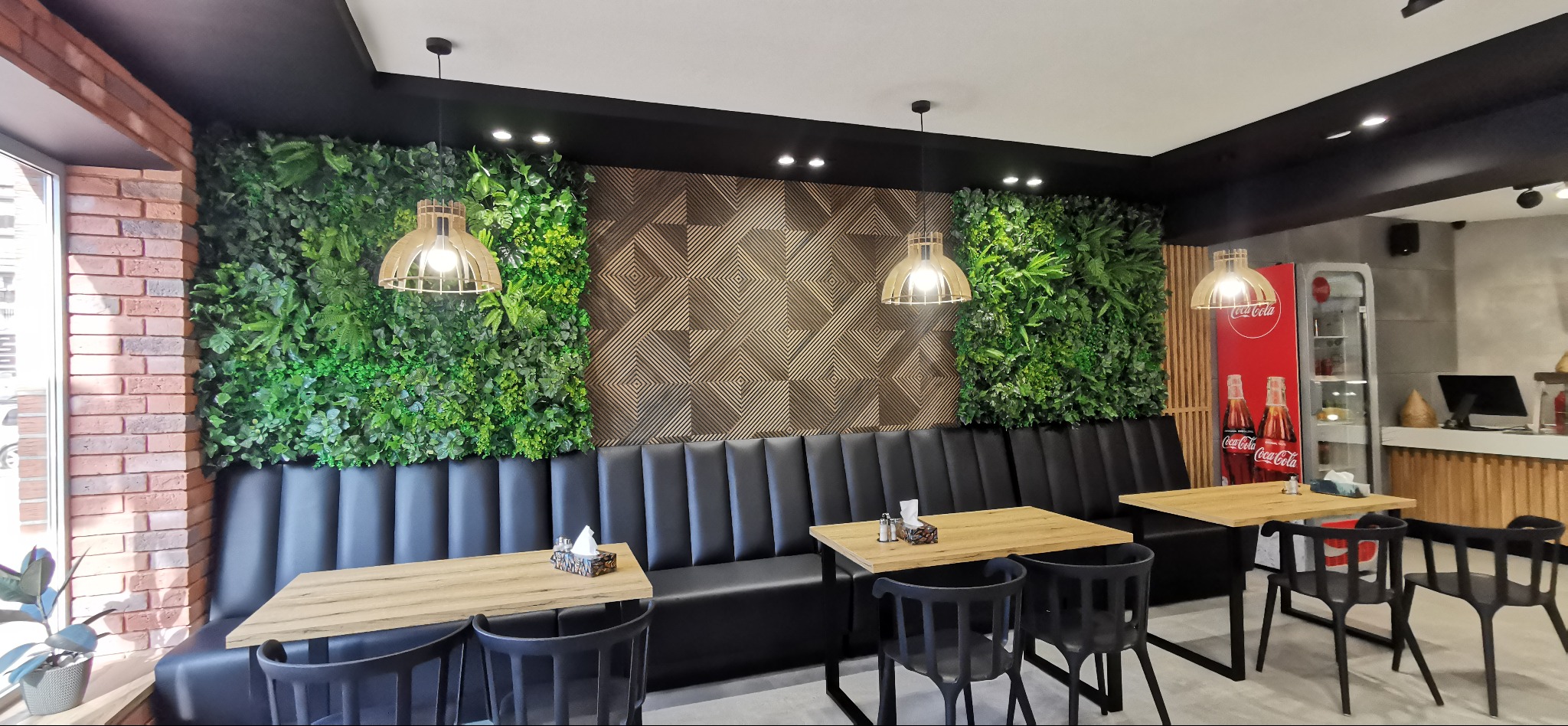 Wnętrze restauracji z zieloną ścianą z roślin, geometrycznym panelem dekoracyjnym, czarną sofą w stylu retro i drewnianymi stolikami pod wiszącymi lampami.