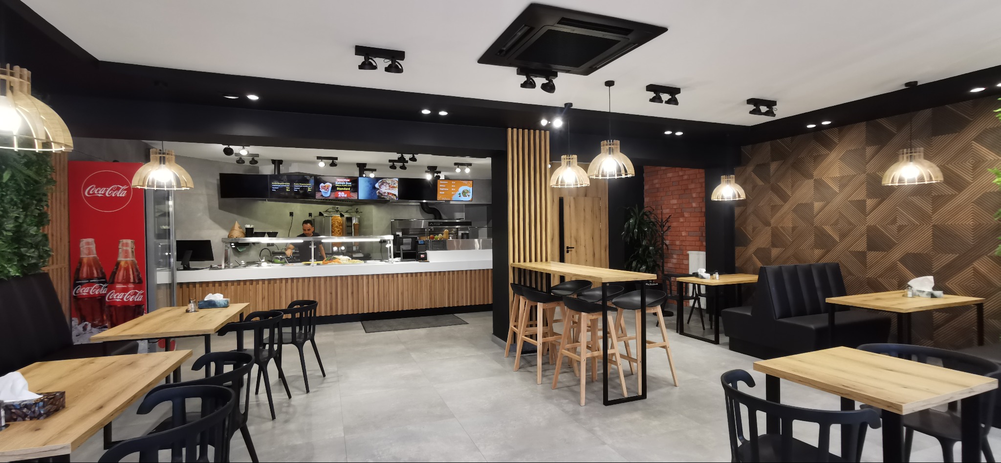 Wnętrze lokalu gastronomicznego z jasnymi drewnianymi stołami i czarnymi krzesłami, oświetlone wiszącymi lampami z drewnianymi abażurami, z widoczną ladą i osobą obsługującą w tle.