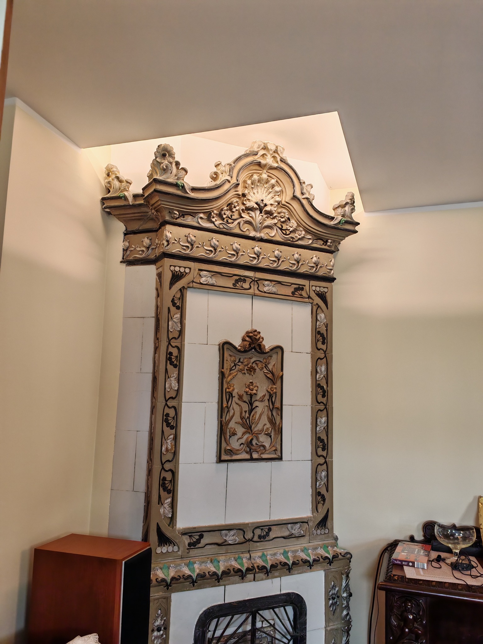 Stary, ozdobny piec kaflowy z misternymi zdobieniami kwiatowymi i zwierzęcymi, umieszczony w rogu pokoju z kremowymi ścianami i oświetleniem sufitowym.