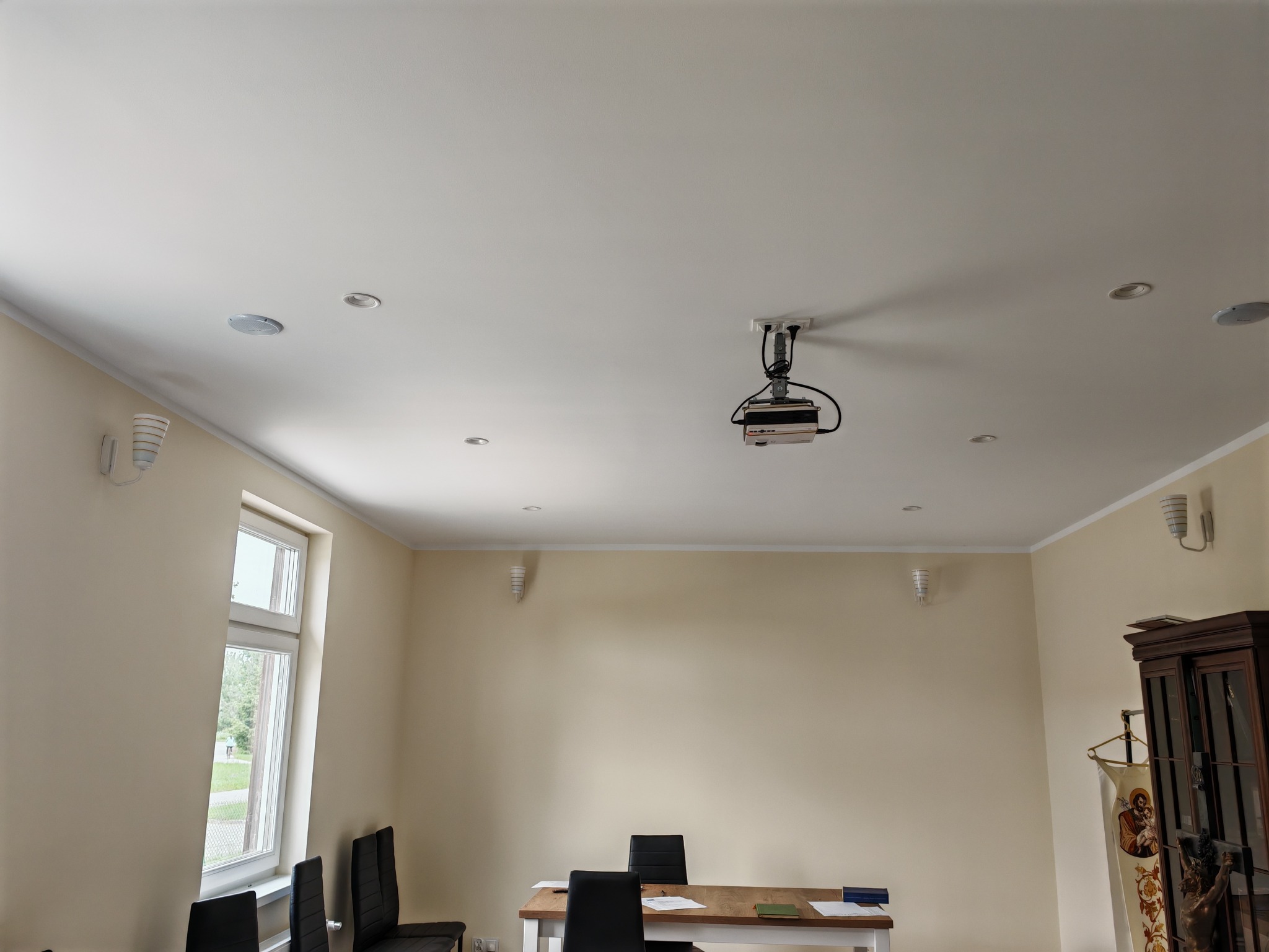 Wnętrze pokoju z zamontowanym projektorem pod sufitem, widoczne także punktowe oświetlenie sufitowe, dekoracyjne kinkiety ścienne, stół z krzesłami oraz szafa w rogu pokoju.