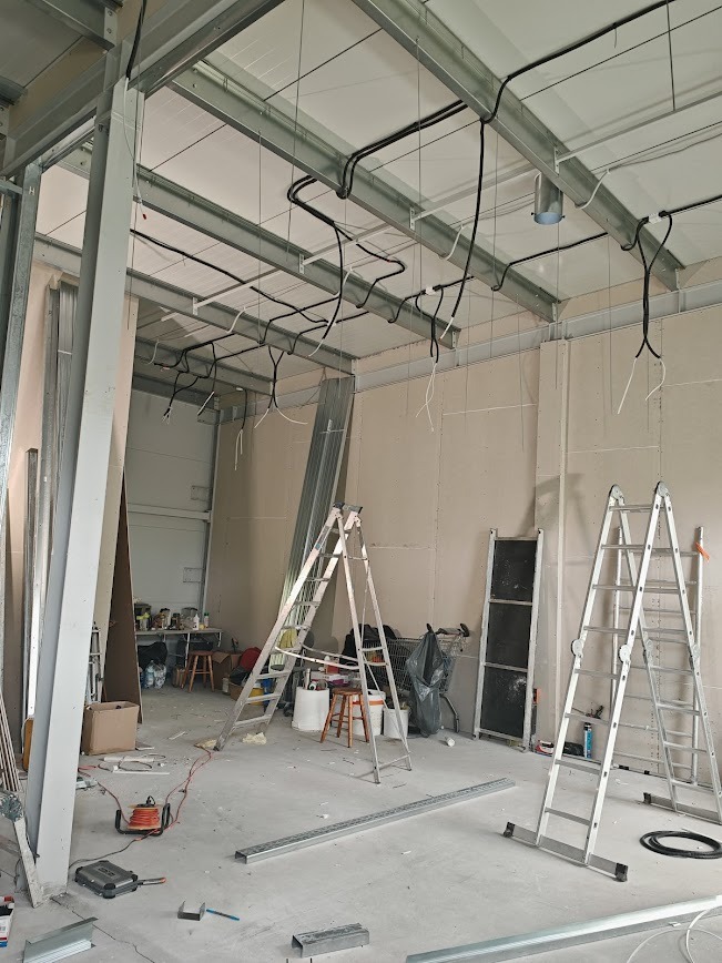 Wnętrze w trakcie prac wykończeniowych: widoczne metalowe profile konstrukcyjne sufitu z poprowadzonymi kablami elektrycznymi, dwie drabiny aluminiowe, narzędzia i materiały budowlane na podłodze.