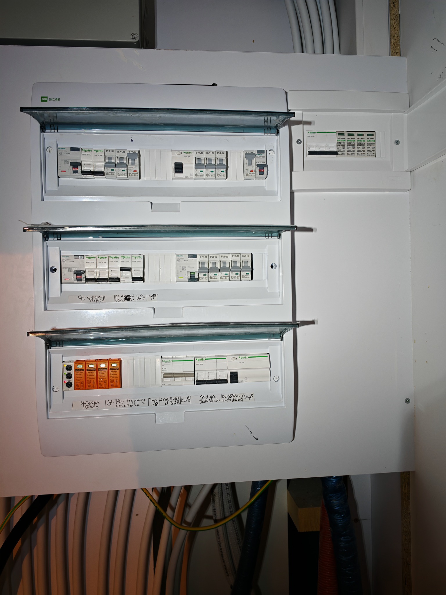 Rozdzielnica elektryczna z bezpiecznikami i opisami, widoczne kable. Instalacja wewnątrz budynku, różne typy zabezpieczeń i marki (Schneider, Eaton).