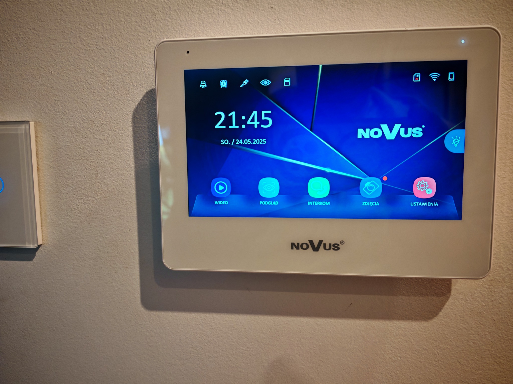 Biały panel dotykowy wideodomofonu Novus zamontowany na ścianie, wyświetlający menu z ikonami i aktualną godziną. Obok widoczny fragment szklanego włącznika.