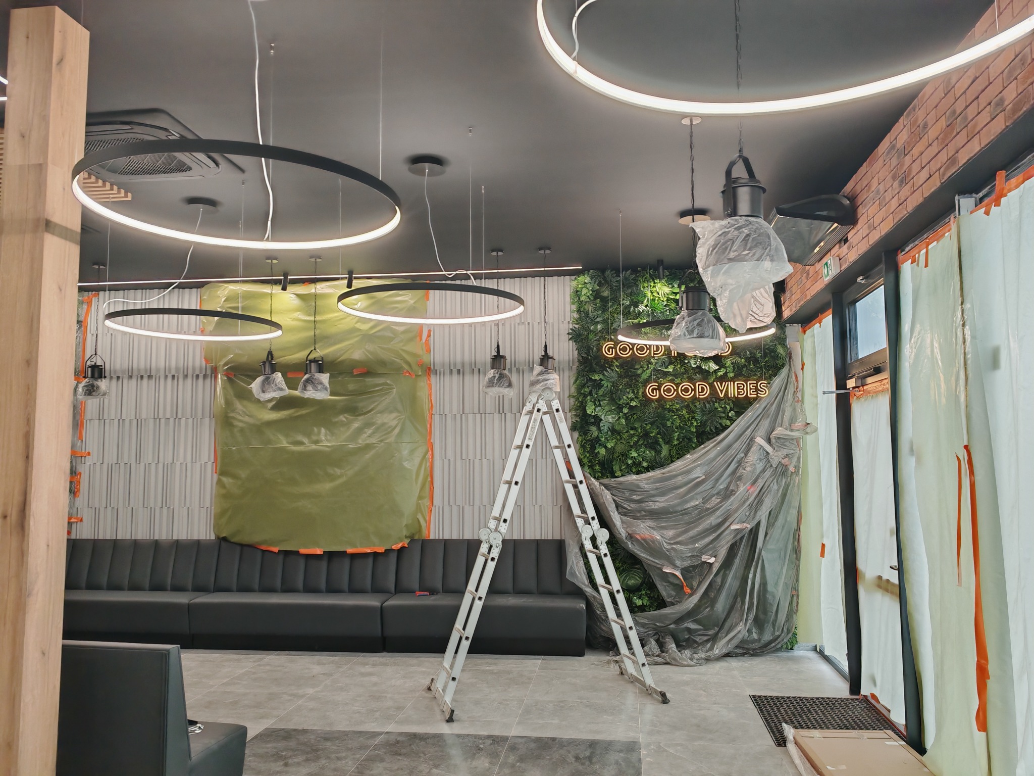 Wnętrze w trakcie montażu oświetlenia: okrągłe lampy LED, drabina, zabezpieczone ściany i meble folią. Widoczny neon 'GOOD VIBES' i fragment ceglanej ściany.