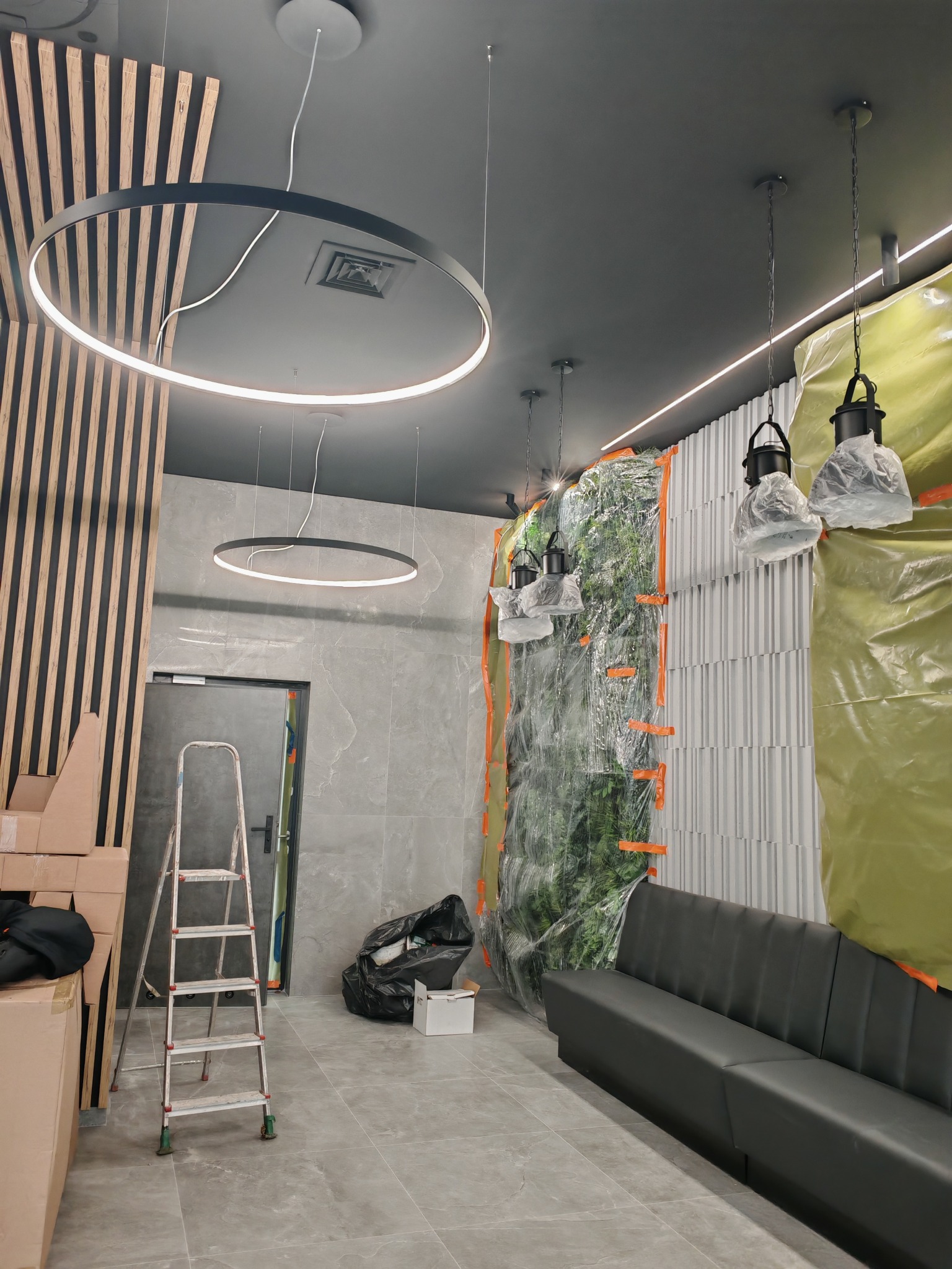 Wnętrze w trakcie montażu oświetlenia: wiszące lampy, okrągłe ledy. Ściana z roślinami zabezpieczona folią. Widoczna drabina i kartony. Szare ściany i sufit.