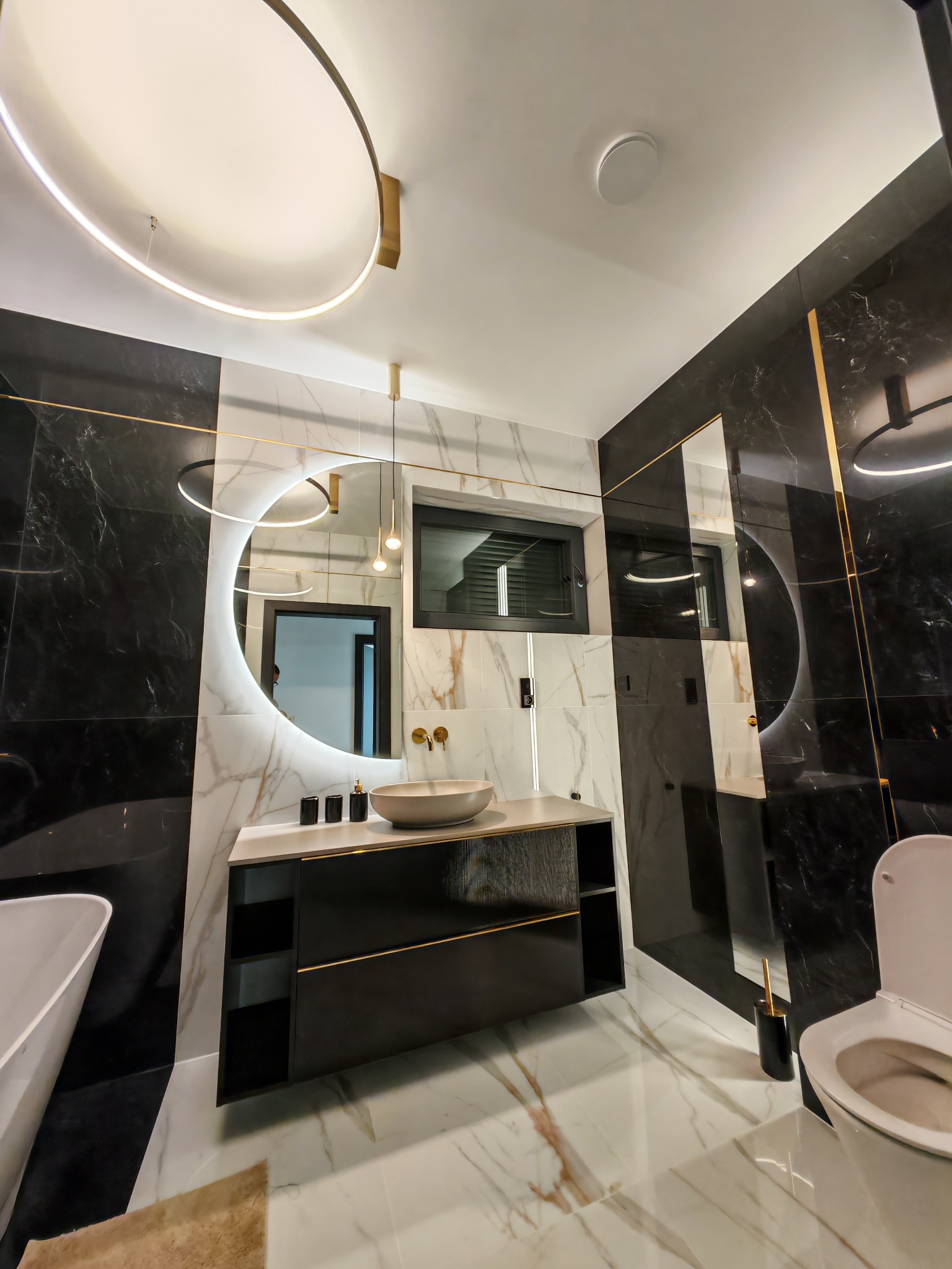 Nowoczesna łazienka z okrągłym lustrem LED, czarnymi i białymi marmurowymi płytkami z złotymi akcentami. Widoczne oświetlenie sufitowe i elementy armatury.