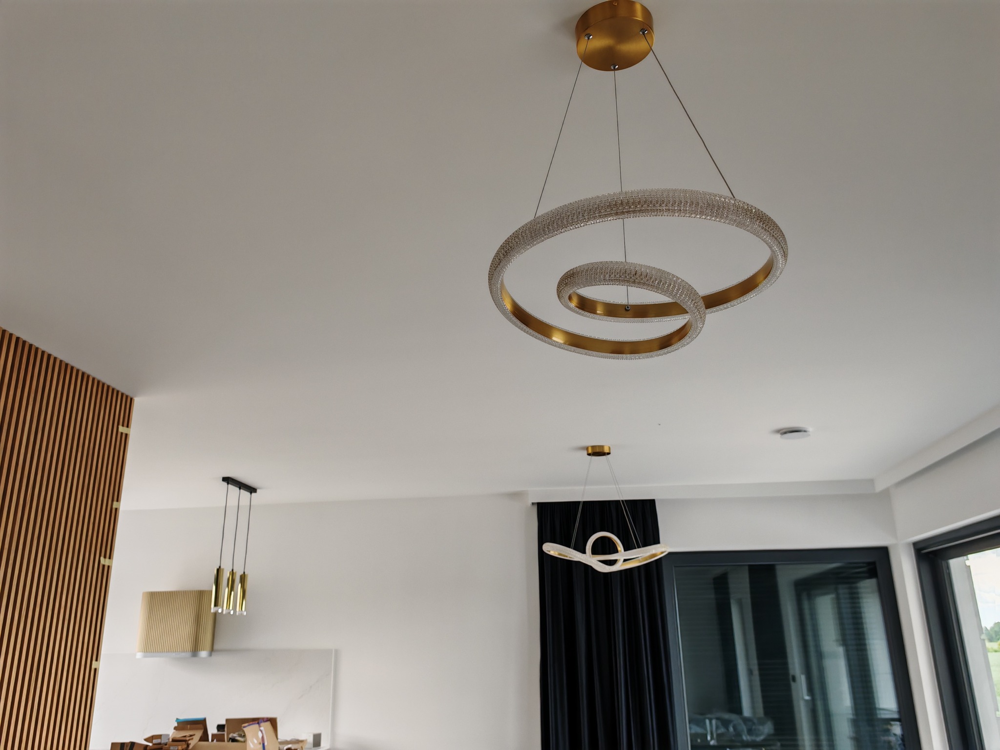 Nowoczesne, złote lampy wiszące w kształcie spiralnych okręgów, oświetlające jasne wnętrze z drewnianą ścianą i oknami. Minimalistyczny design, eleganckie wykończenie.