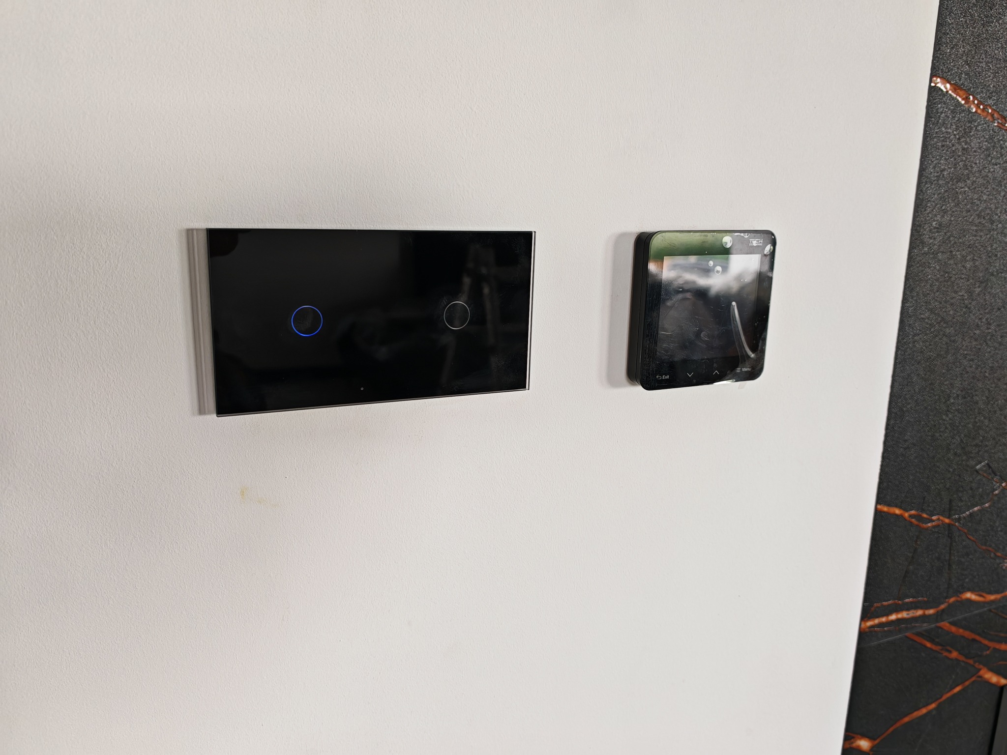 Nowoczesny, dotykowy włącznik światła i panel sterowania domem inteligentnym zamontowane na białej ścianie, obok fragmentu dekoracyjnej płyty.