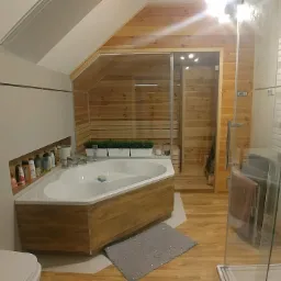 Nowoczesna łazienka na poddaszu z drewnianymi panelami, narożną wanną obudowaną drewnem i szklaną kabiną prysznicową.