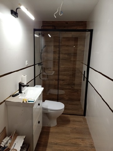 Nowoczesna łazienka w trakcie wykańczania, widoczna kabina prysznicowa z czarnymi ramami, biała umywalka z czarną armaturą, toaleta oraz płytki imitujące drewno na podłodze i ścianie prysznica...